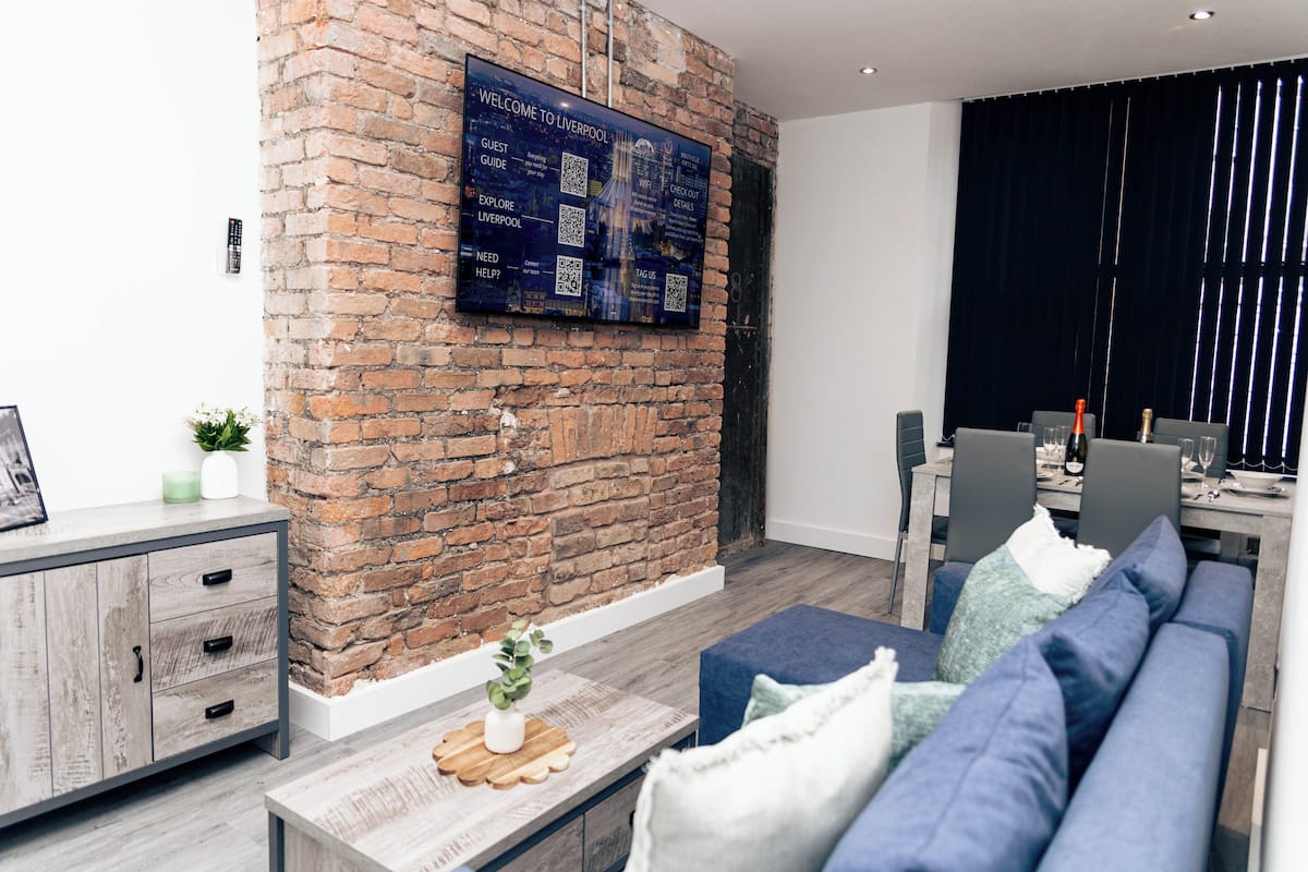 A modern living room with a brick wall, a mounted screen displaying information, a dining area with chairs and a table, and a sofa with cushions.