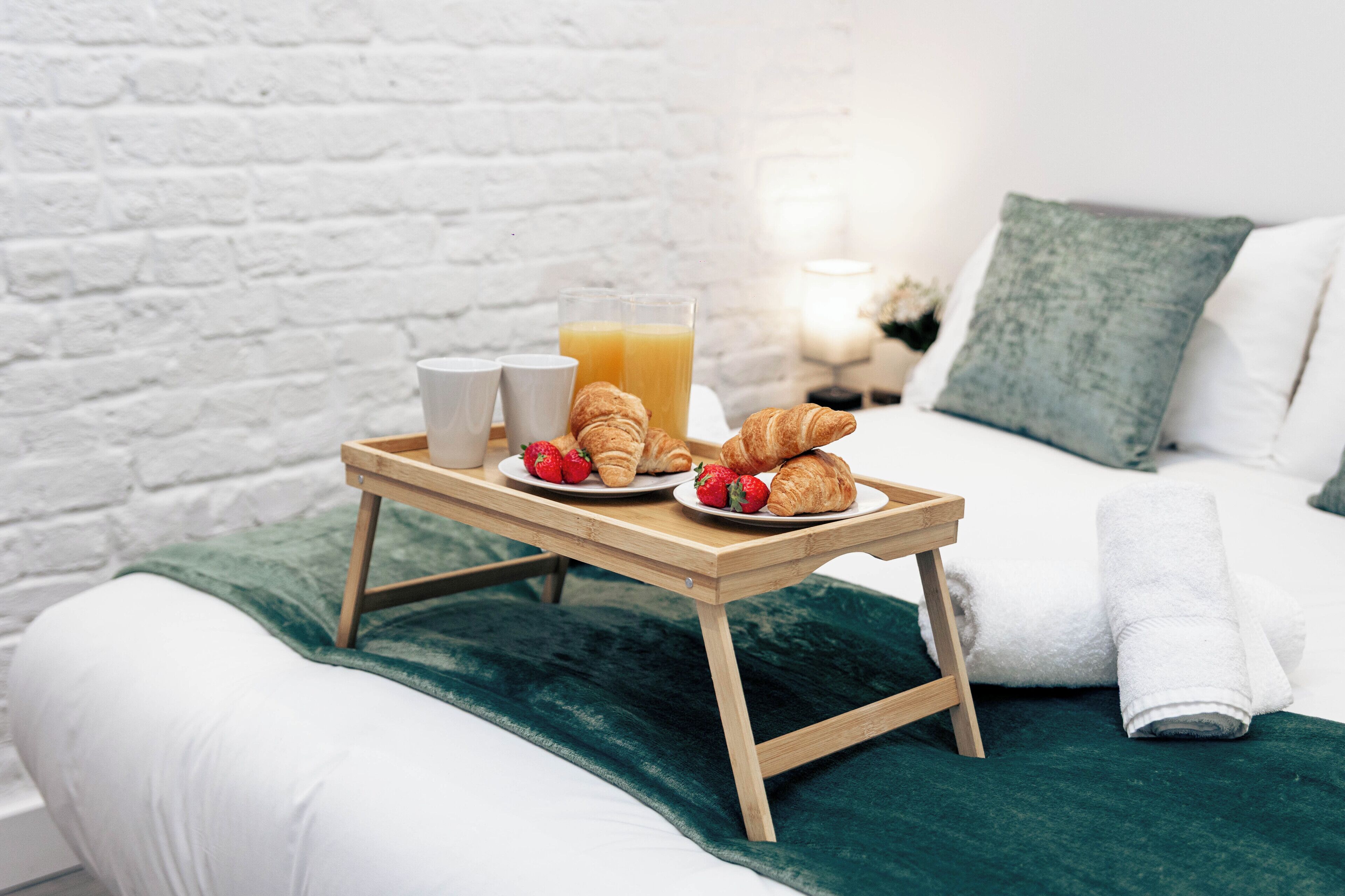 Un letto con una coperta verde, un comodino in legno con prodotti per la colazione, e un muro di mattoni bianchi.