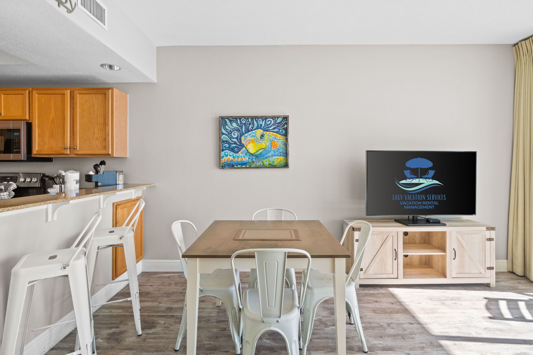 A modern living room with a wooden dining table, white chairs, a flat-screen TV, and a painting on the wall.
