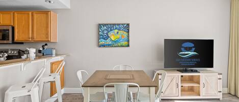 A modern living room with a wooden dining table, white chairs, a flat-screen TV, and a painting on the wall.