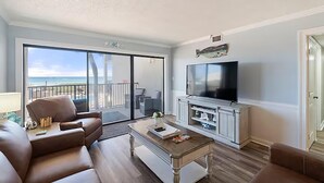 Una sala de estar con televisor grande, sofá, mesa de centro y balcón con vista al océano.