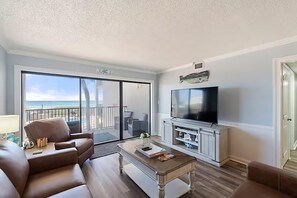 A living room with a large TV, a sofa, a coffee table, and a balcony with a view of the ocean. - Moondrifter 205 (Panama City Beach)