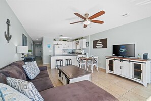 A modern living room with a brown sofa, a wooden coffee table, and a ceiling fan. There is a kitchen area with white cabinets and a TV on a wooden stand. - Beach Front, “Wave N' Sea” 202, Free Activities Included! (Fort Walton Beach)
