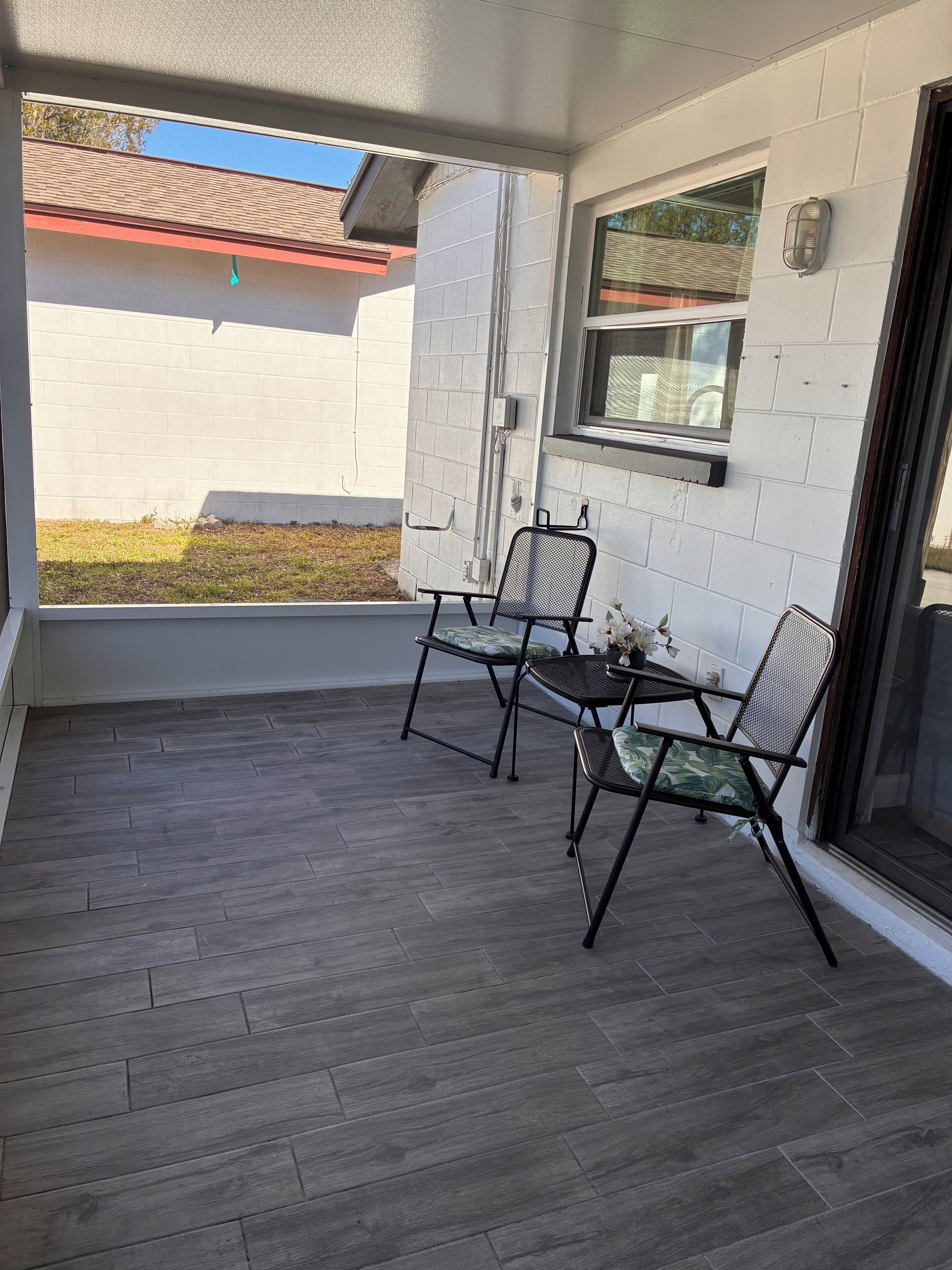 Une terrasse couverte avec une table et deux chaises, une fenêtre et une vue sur un immeuble voisin.