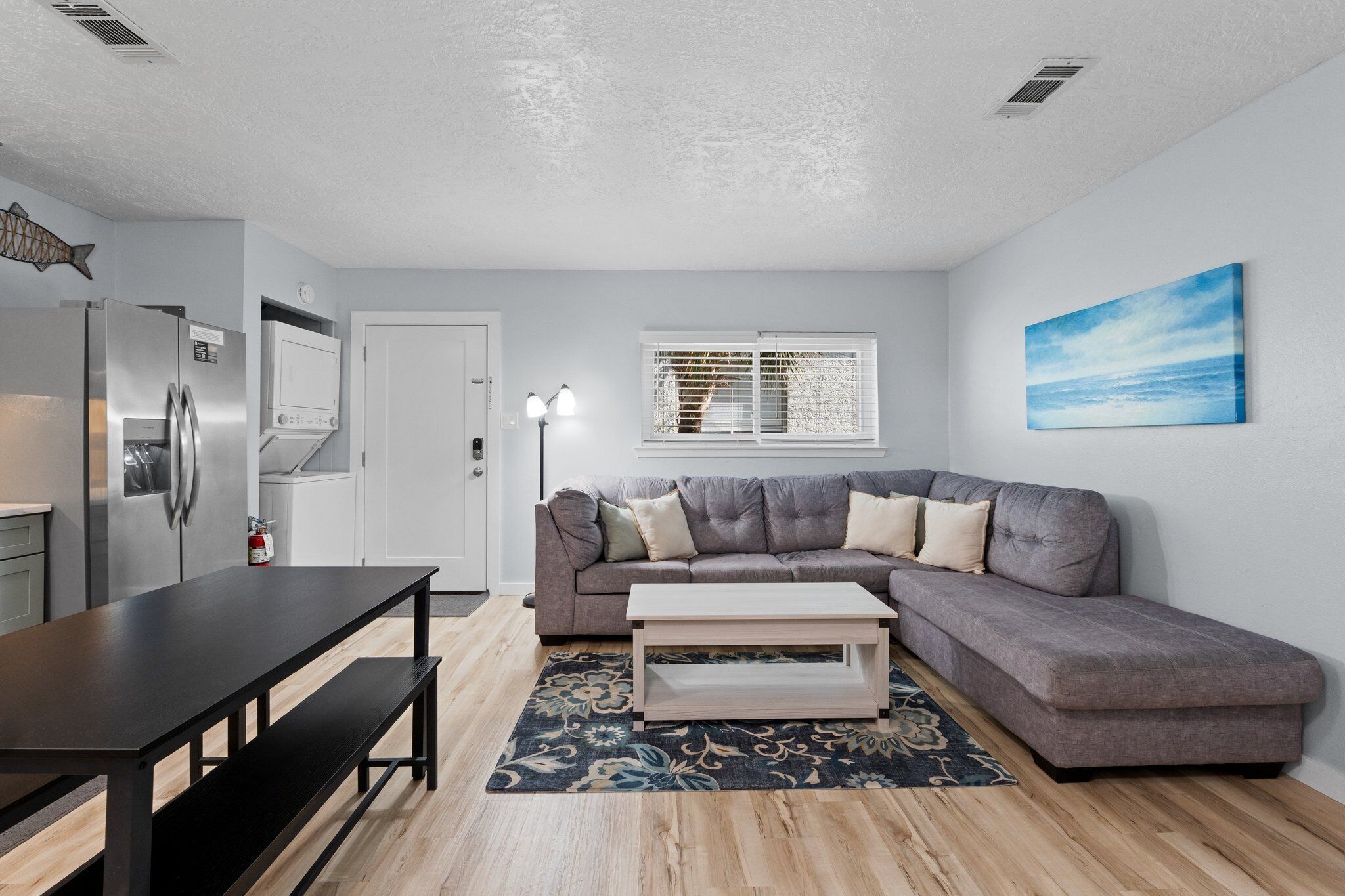 A modern living room with a gray sectional sofa, a wooden coffee table, a dark patterned rug, and a kitchen area with a stainless steel refrigerator.