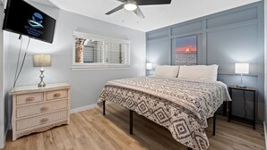 Ein Schlafzimmer mit einem Bett, Deckenventilator, Fernseher, Fenster mit Jalousien und einem Wandbild.