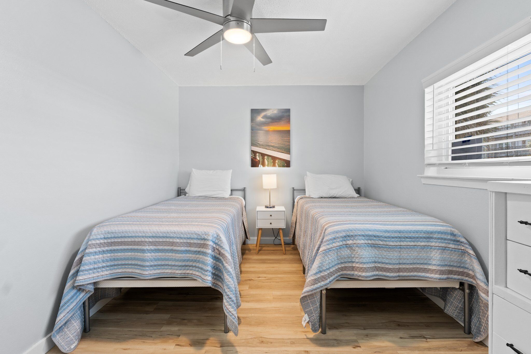 A bedroom with two single beds, a ceiling fan, a nightstand with a lamp, and a window with blinds.