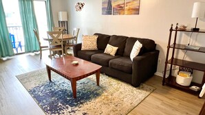 Un soggiorno con un divano marrone, un tavolino in legno, un tappeto blu e beige, una zona pranzo con sedie, una lampada e una libreria.