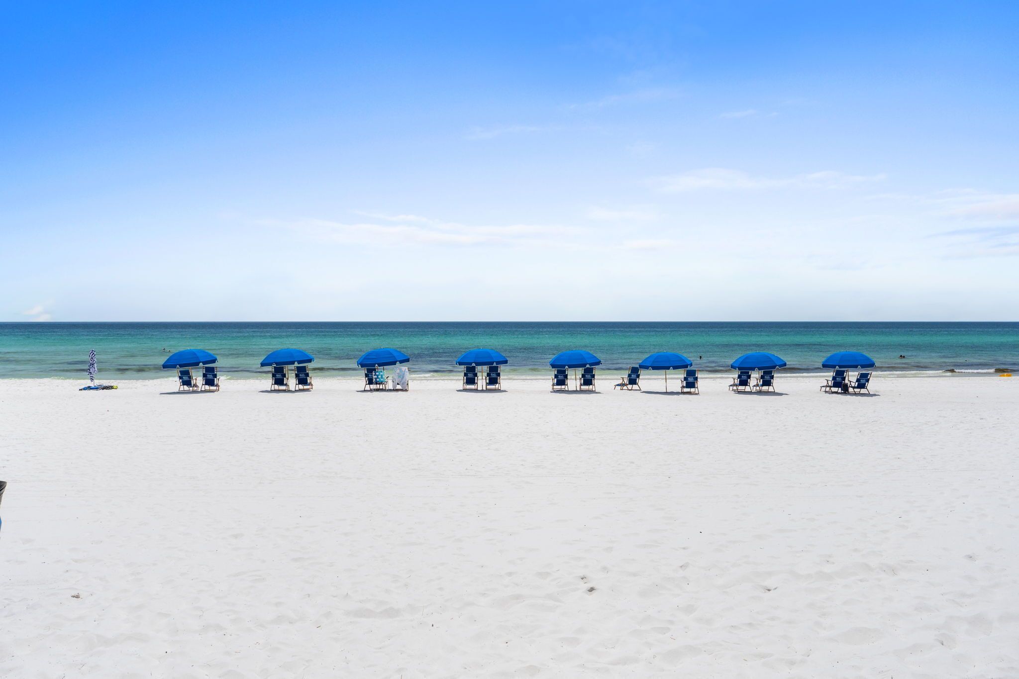 A beach with white sand and a clear blue sky. There are blue umbrellas and lounge chairs arranged in a row.