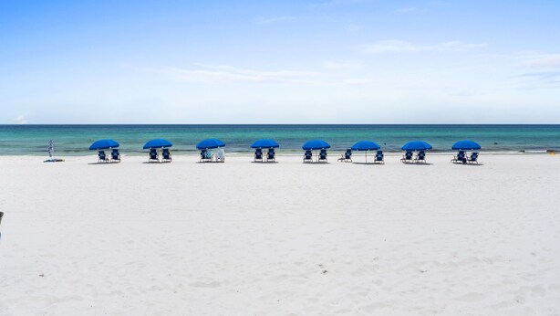 Une plage de sable blanc, sous un ciel bleu azur. Des parasols bleus et des chaises longues sont disposés en rangée.