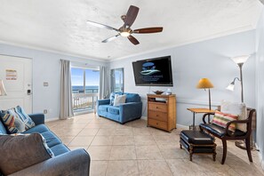 A spacious living room with a blue sofa, a wooden chair, a small table, and a ceiling fan. A television is mounted on the wall displaying a logo. - Beach Service~Dog Friendly~Gulf Front Condo~Pool~Free Activities (Fort Walton Beach)