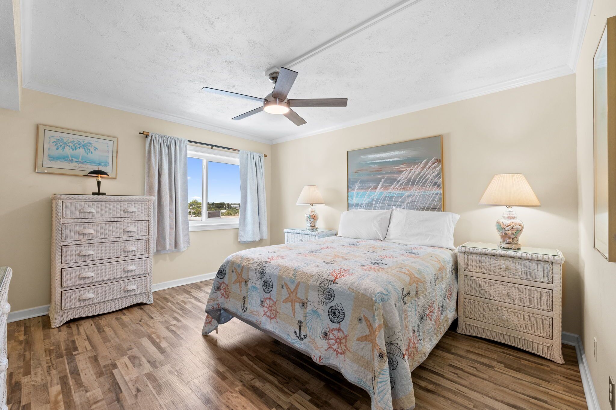 Une chambre à coucher comprenant un lit, des tables de chevet, une commode, un ventilateur de plafond et une fenêtre avec des rideaux.