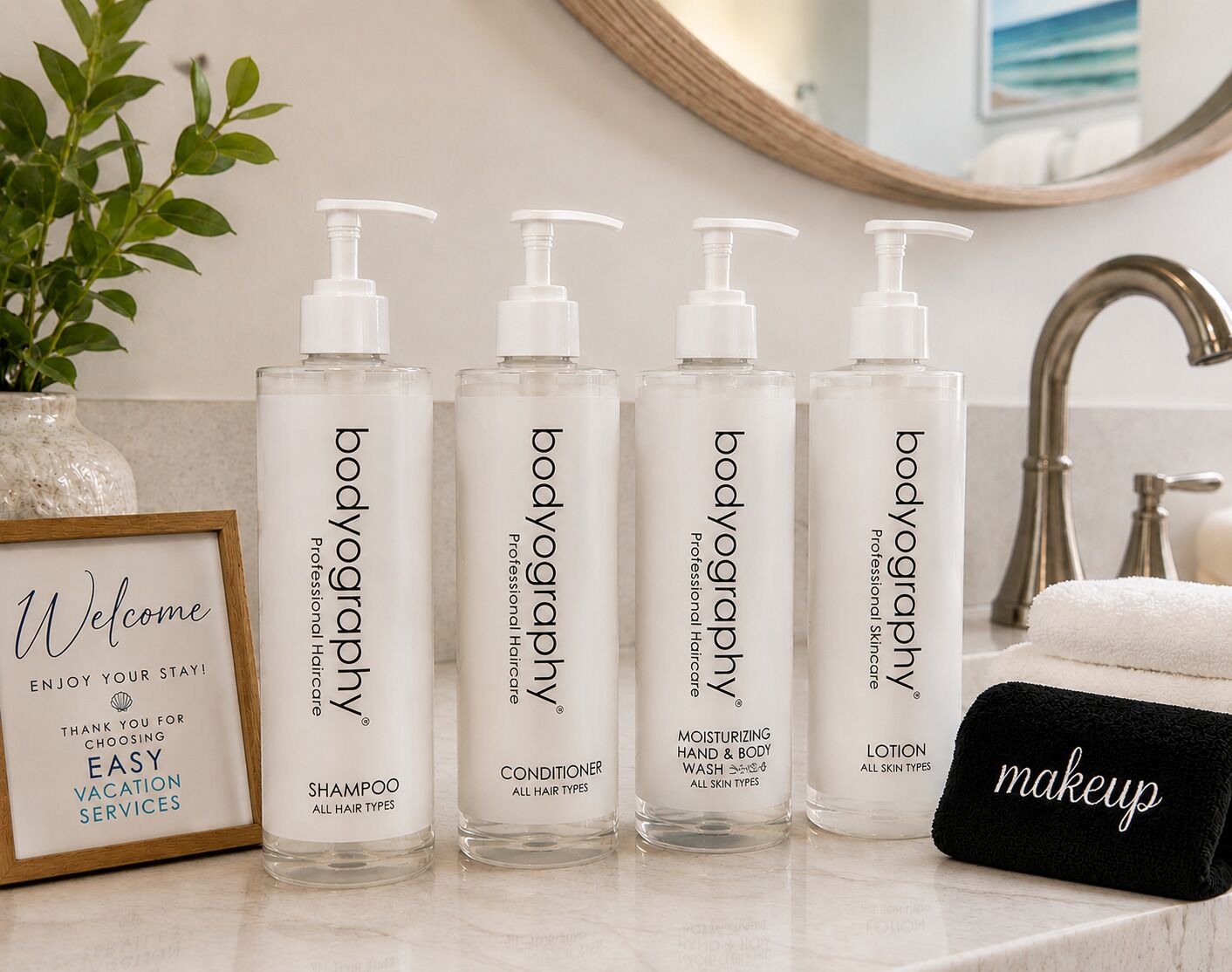 Four bottles of bodyography products, a framed welcome sign, and a black makeup bag on a bathroom counter.