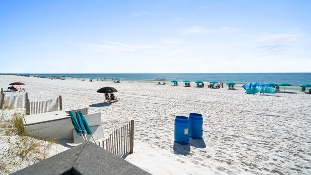 Ein Sandstrand mit Sonnenschirmen und Liegestühlen, eine Holzlattenveranda und ein klarer, blauer Himmel.