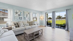 Een woonkamer met een bank, salontafel en kunstwerk aan de muur. Er is een raam met gordijnen, een lamp en uitzicht op het landschap.