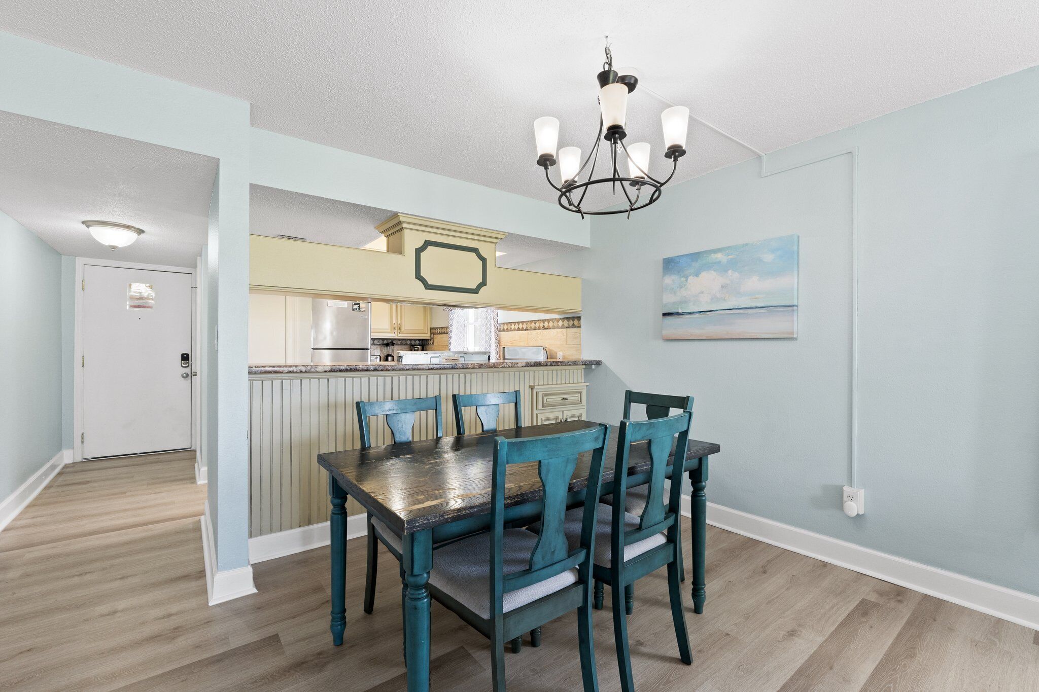 A dining area with a wooden table and chairs, a kitchen in the background, and a light blue wall with a painting.