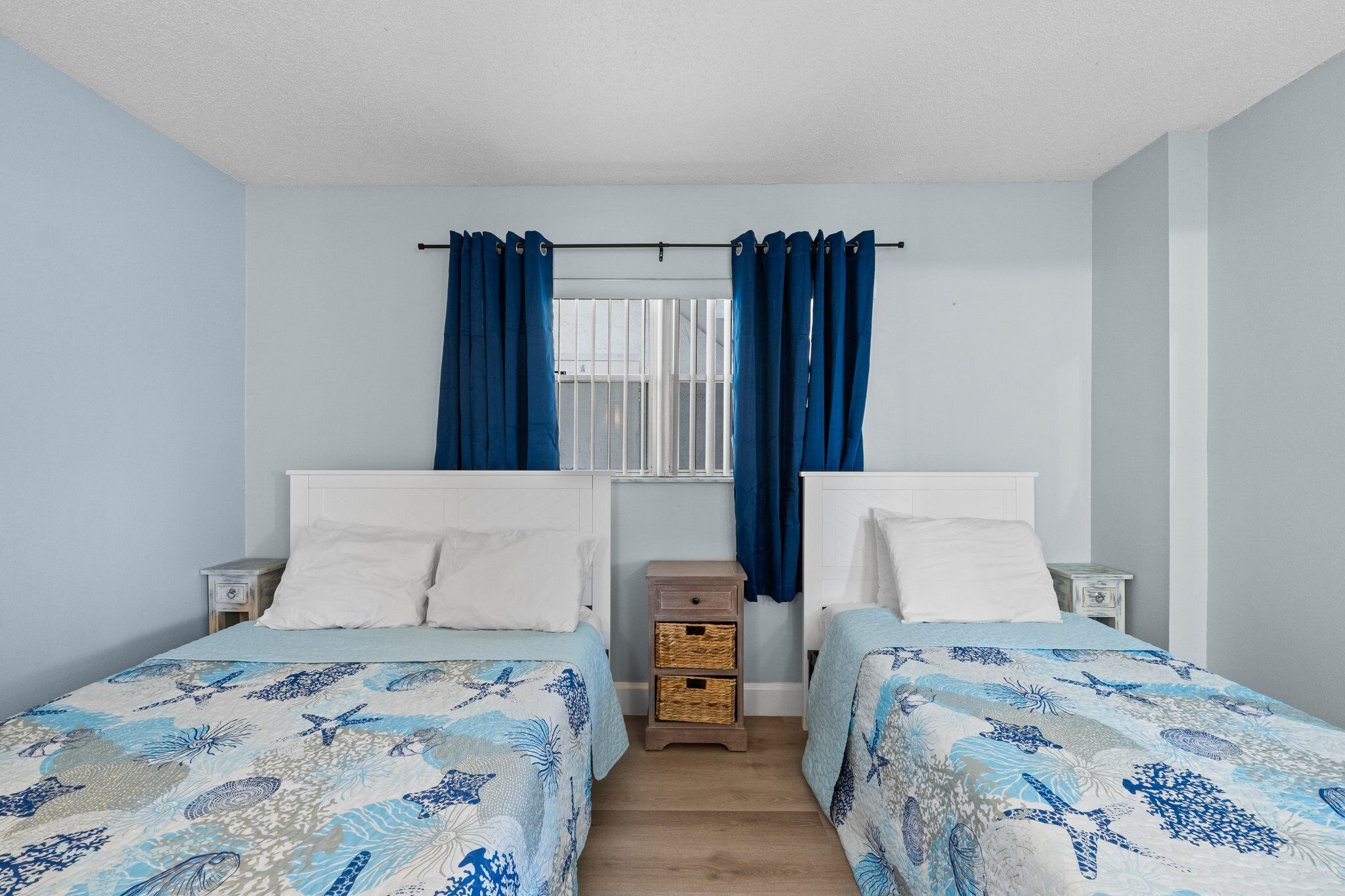 Un dormitorio con dos camas, una ventana con cortinas, un pequeño aparador de madera y un cabecero blanco.