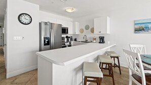 A modern kitchen with white cabinetry, a stainless steel refrigerator, a microwave, and a sink. There are two bar stools and a dining table with chairs. A clock is mounted on the wall, and a painting of chairs on the beach is visible. - New! Sea La Vie: Beachfront Bliss at The Veranda (Fort Walton Beach)