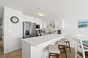 A modern kitchen with white cabinetry, a stainless steel refrigerator, a microwave, and a sink. There are two bar stools and a dining table with chairs. A clock is mounted on the wall, and a painting of chairs on the beach is visible.