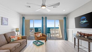 En moderne stue med sofa, sofabord og fjernsyn på væggen. Der er en balkon med udsigt til stranden og en ventilator i loftet.
