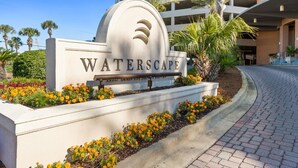 Letrero de 'WATERSCAPE' con diseño moderno, rodeado por un jardín ajardinado con flores amarillas y palmeras.