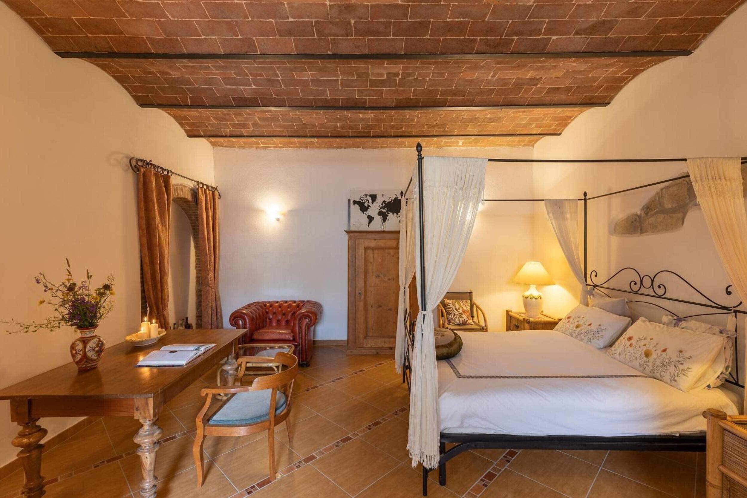 Una camera da letto con un letto a baldacchino, una scrivania in legno con una sedia, una poltrona rossa e un tavolino con un vaso e delle candele.