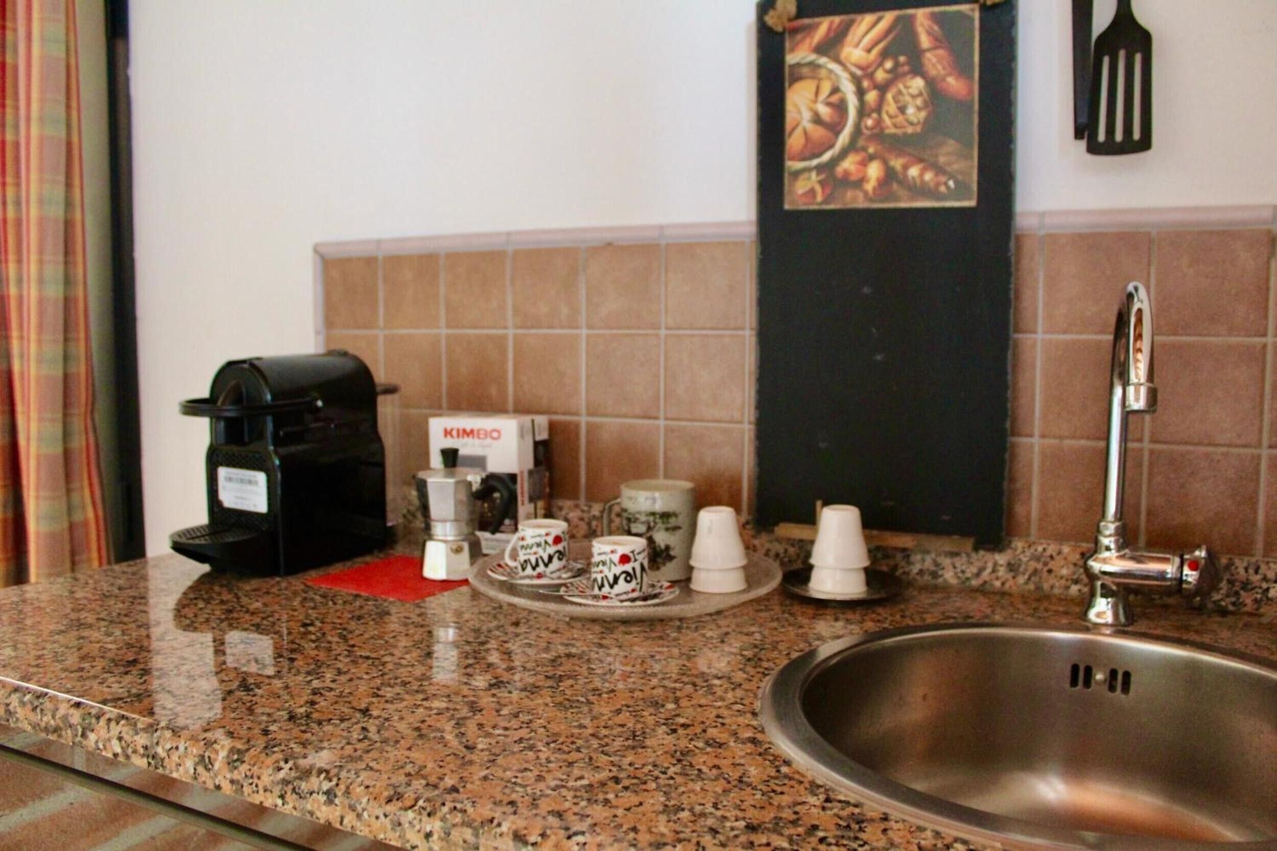 A kitchen with a granite countertop, a coffee maker, disposable cups, a sink, and a spatula on the wall.