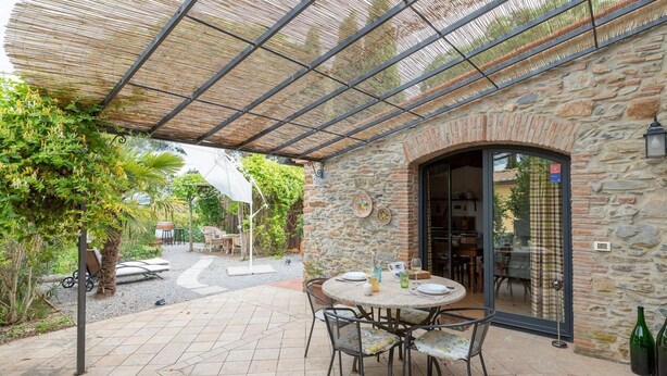 Un patio al aire libre con un muro de piedra, una mesa redonda con sillas y una zona cubierta con techo de metal.