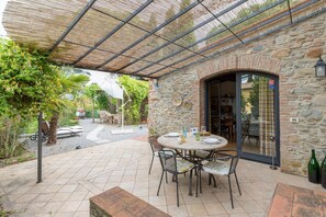 En udendørs terrasse med en stenvæg, et rundt bord med stole og et overdækket område med et metal tag.