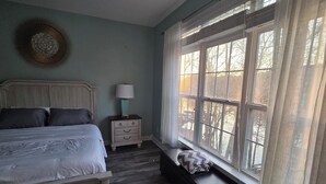 A bedroom with a bed, a nightstand, a lamp, a bench by the window, and a view of trees outside.
