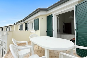 Ein Balkon mit einem weißen Tisch und Stühlen, einem Bett mit grünen Rollläden und einem weißen Geländer.