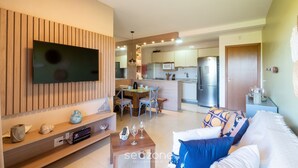 A modern living room with a flat-screen TV, wooden panel wall, and a dining area with a kitchen in the background. - Apt in Condo w/ Pool 750 m Beach Rdw0103 (Camaçari)