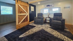 Een kamer met een deur in schuurstijl, twee grijze fauteuils, een vloerkleed met dessin, een lijstwerk en een bord aan de muur.