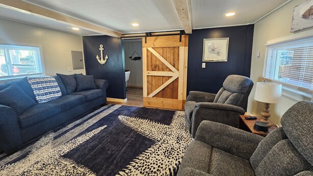 Een gezellige woonkamer met een donkerblauwe bank, een grijze fauteuil en een kamerkleed met een patroon. Op de achtergrond is een houten deur met een boerderij-stijl zichtbaar.