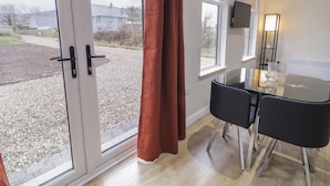 A room with a glass door leading to a gravel area, a table with chairs, a TV mounted on the wall, and a floor lamp.