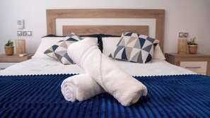 Ein ordentlich bezogenes Bett mit einer blauen, strukturierten Tagesdecke, zwei Kissen mit geometrischen Mustern und einem aufgerollten weißen Handtuch.