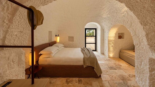 Une chambre avec un mur en pierre, un lit, une table de chevet et une fenêtre.