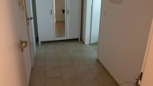En hall med ett kaklat golv, vita väggar och en taklampa. Det finns två dörrar på höger sida och en liten vit stol i förgrunden.