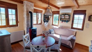 A cozy room with a wooden ceiling, a round wooden table with chairs, a sofa, a radiator, a stove, and a window with a view of greenery.