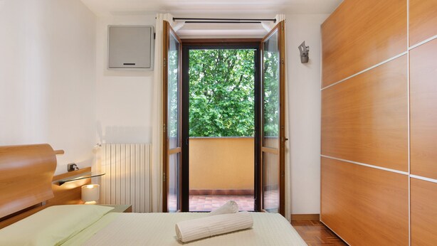 A hotel room with a bed, a wooden wardrobe, a radiator, and a view of greenery through an open door.