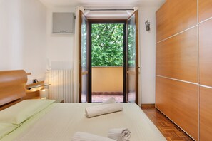 Quarto de hotel com cama, guarda-roupa de madeira, radiador e vista para o verde através de uma porta aberta.
