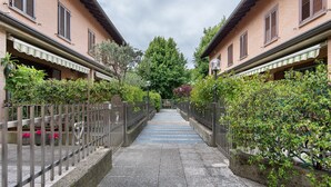 A narrow pathway flanked by buildings with balconies, a metal fence, and greenery.