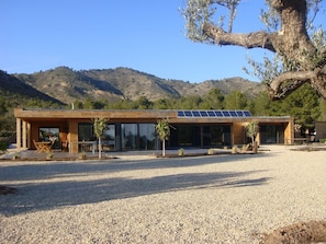 Un edificio moderno con camino de grava, paneles solares y vistas a las montañas.