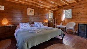Een slaapkamer in een houten hut met een bed, twee bedlampen, een stoel, een open haard en een raam.