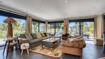 A spacious living room with a sofa, armchair, coffee table, and a view of the outdoors through large windows.