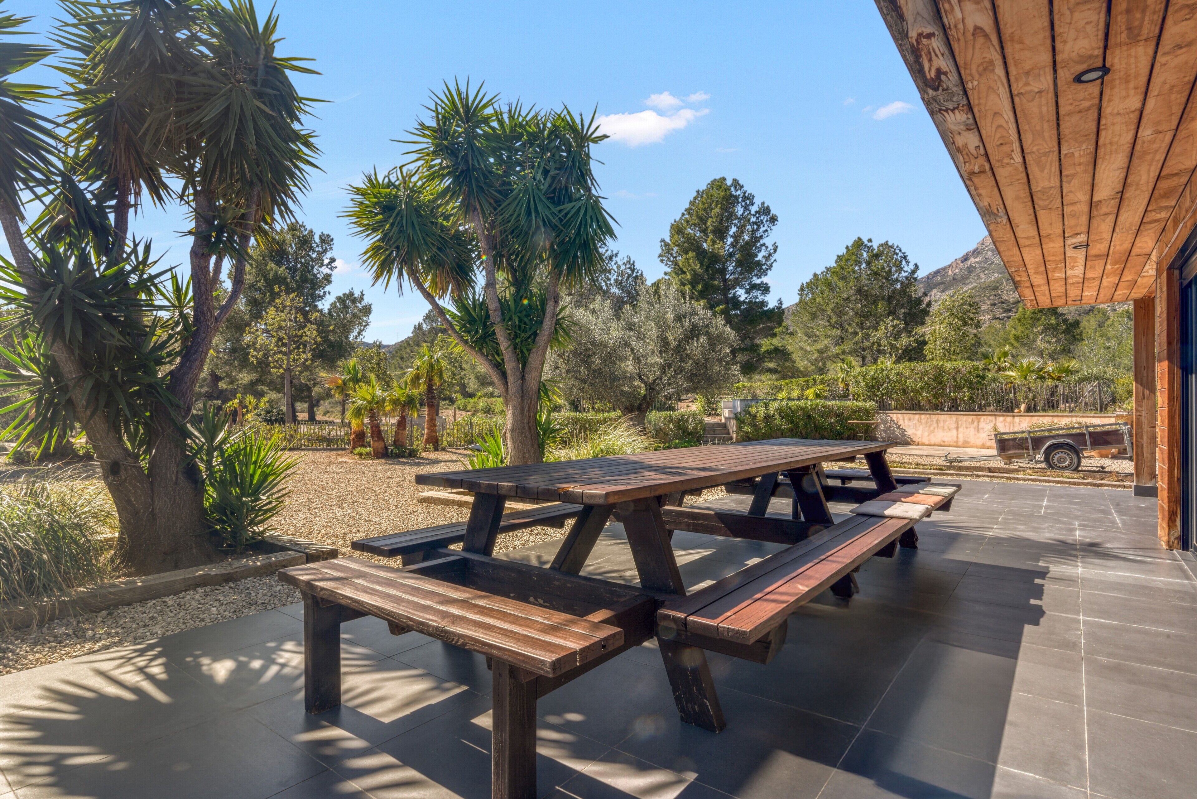 A patio with wooden picnic tables, surrounded by trees and a clear sky.