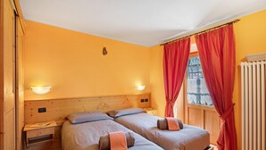 Quarto com duas camas, cabeceira de madeira, mesas de cabeceira, luminárias fixadas na parede, janela com cortinas vermelhas e um radiador.