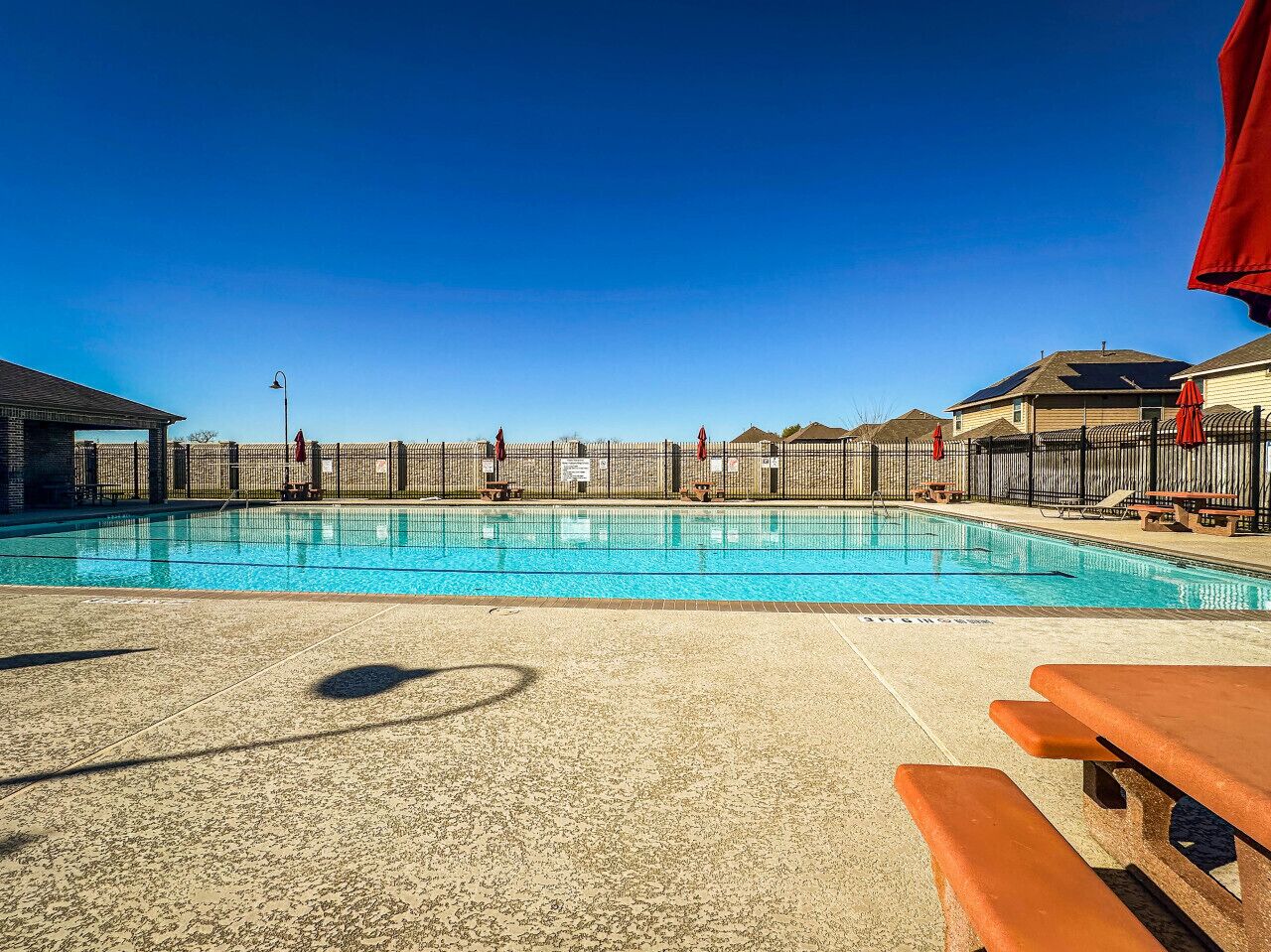 A swimming pool with a clear blue sky, a fence, and picnic tables.
