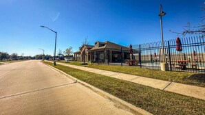 A clear road with a sidewalk, a building with a fenced pool area, and a clear blue sky. - Alpha Group - Room 1 (Richmond)