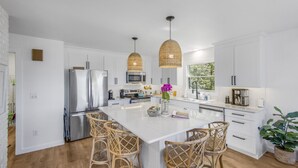 Een moderne keuken met witte kasten, roestvrijstalen apparatuur, rieten hanglampen en een centraal eiland met barkrukken.
